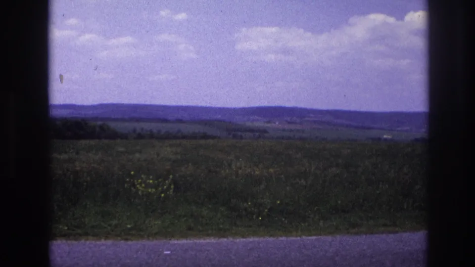 8mm home movie, France c.1967 — wide rural landscape with rolling green fields, distant wooded hills, and a large purple-tinged sky with scattered white clouds.