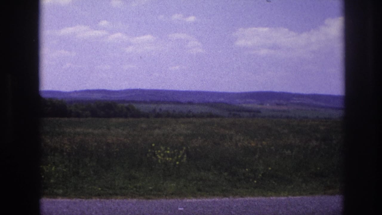 8mm home movie, France c.1967 — open agricultural countryside with fields, wooded tree line, low hills, and a purple-tinted sky in a nearly unchanged composition.