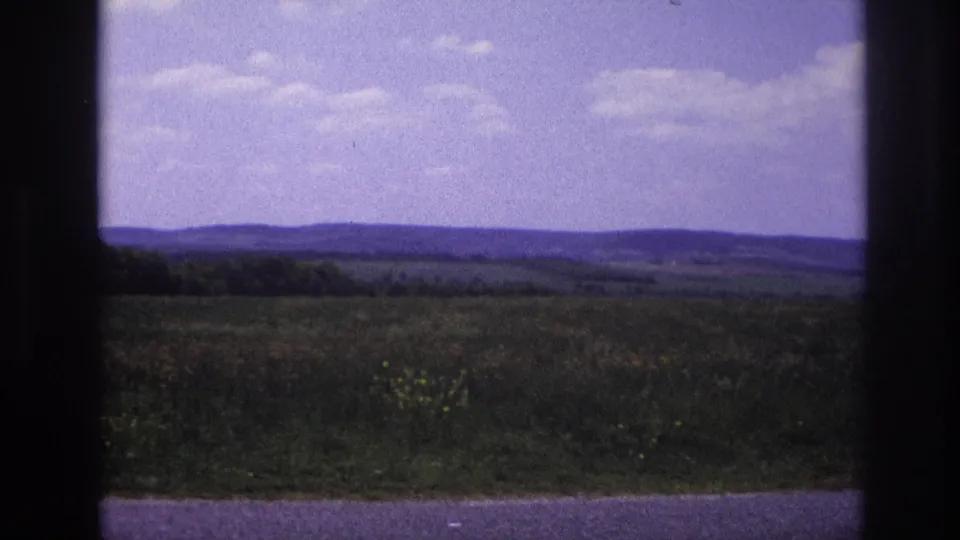 8mm home movie, France c.1967 — open agricultural countryside with fields, wooded tree line, low hills, and a purple-tinted sky in a nearly unchanged composition.