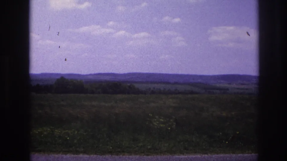 8mm home movie, France c.1967 — wide-angle view with sky dominating the upper half, rolling green countryside below, and visible film dust artifacts throughout.