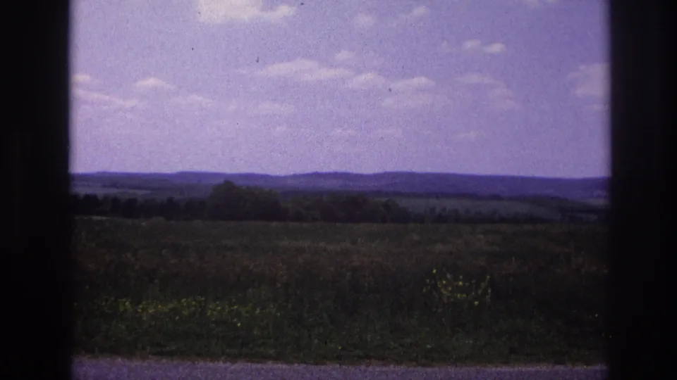 8mm home movie, France c.1967 — final frame showing wide countryside with yellow wildflowers in lower right, tree line, distant hills, and a purple-cast sky with cumulus clouds.