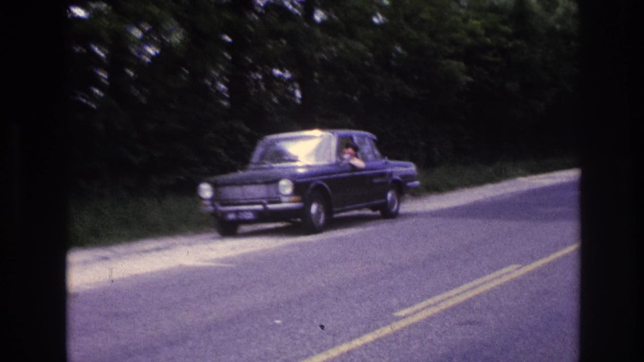Dark blue European compact sedan on a tree-lined two-lane road in France, circa 1967; driver partially visible through windshield, yellow road markings visible.