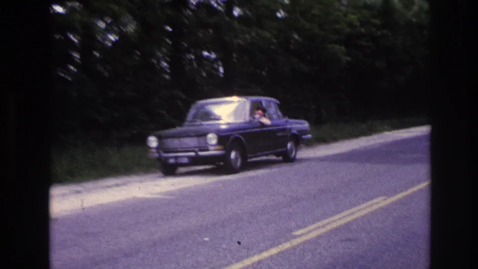 Dark blue European compact sedan on a tree-lined two-lane road in France, circa 1967; driver partially visible through windshield, yellow road markings visible.