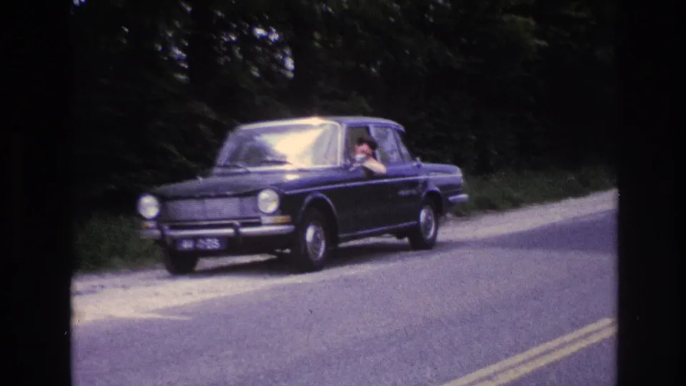 Close view of dark blue European compact sedan with horizontal ribbed grille and round headlights on a French rural road, circa 1967; person visible at driver's window.