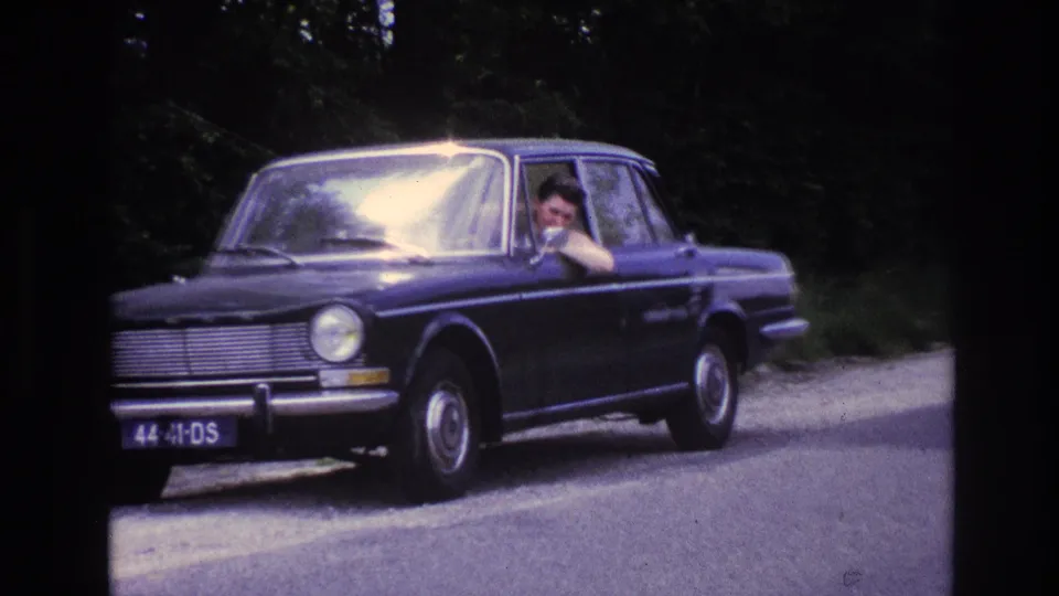 Close front view of dark blue European compact sedan in France, circa 1967; license plate '44 41 DS' clearly legible, driver visible at window, tree-lined road behind.