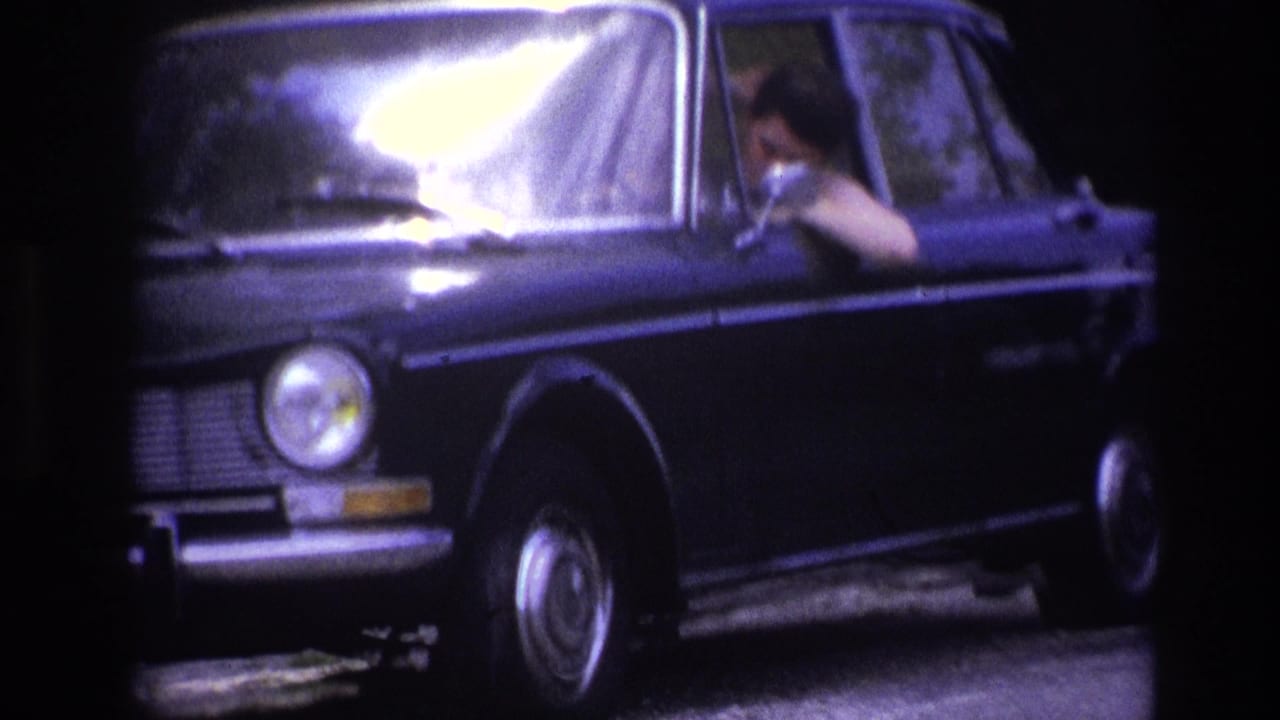 Front-right close shot of a 1960s dark blue European sedan on a French rural road, driver glimpsed through bright windshield glare, dense trees behind.