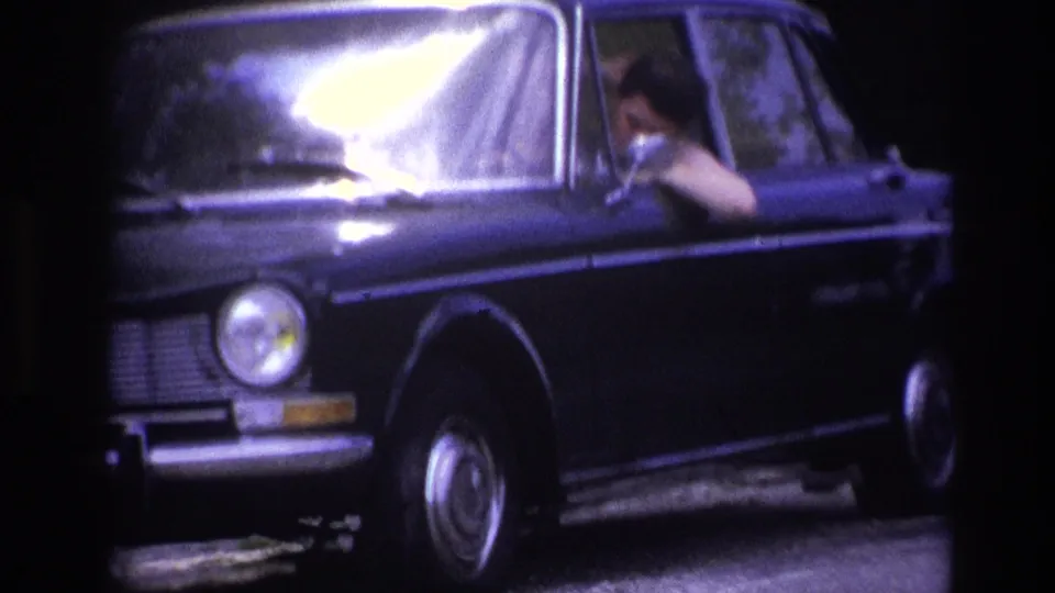 Front-right close shot of a 1960s dark blue European sedan on a French rural road, driver glimpsed through bright windshield glare, dense trees behind.