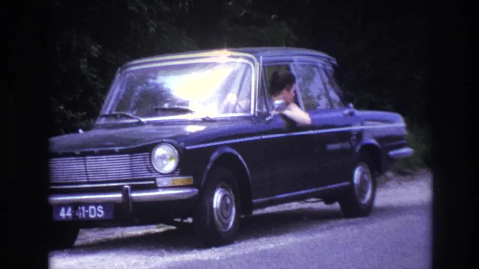 1960s dark blue European sedan stopped on a French rural road, driver with arm raised at open window, French license plate characters legible on front bumper.