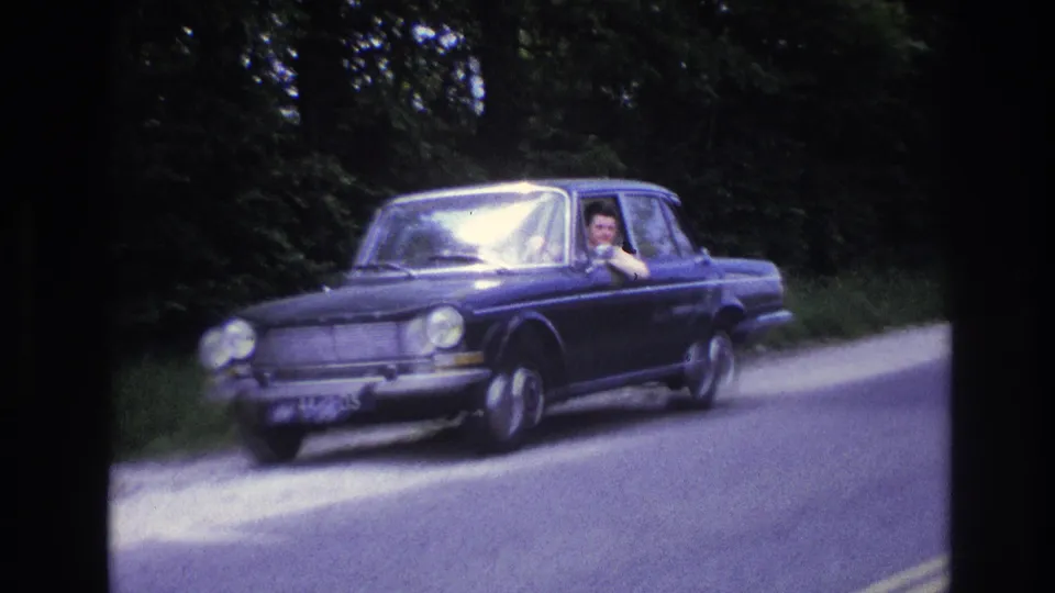 Wide shot of a 1960s dark blue European sedan on a tree-lined rural French road, center lane marking visible on paved surface, driver seen through windshield.