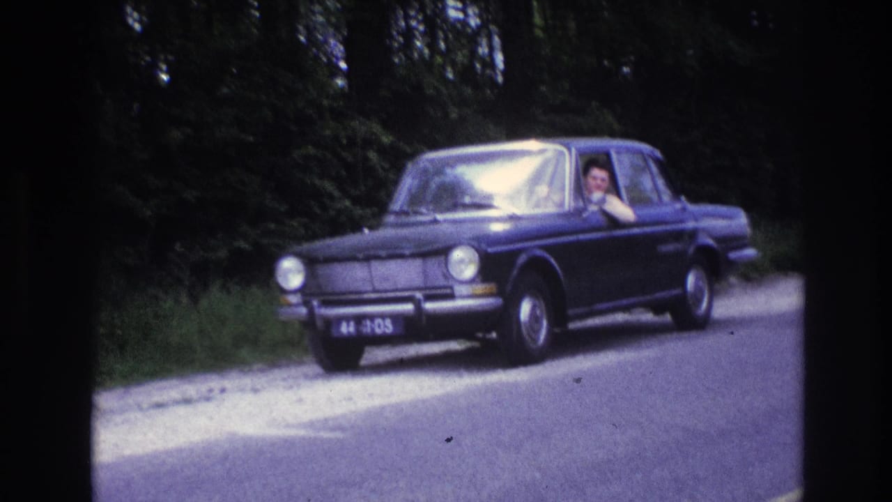Mid-1960s dark blue sedan on a French rural road, circa 1967; the driver is visible waving through the open window with a dense green treeline behind.