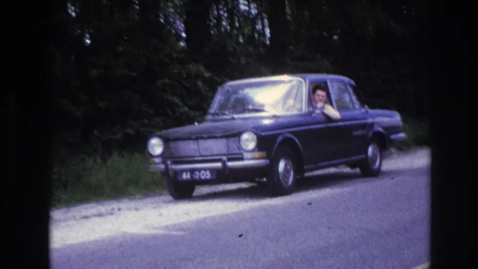 Mid-1960s dark blue sedan on a French rural road, circa 1967; the driver is visible waving through the open window with a dense green treeline behind.