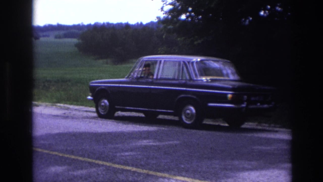 Dark blue sedan viewed from behind driving away on a rural road, open green fields visible.
