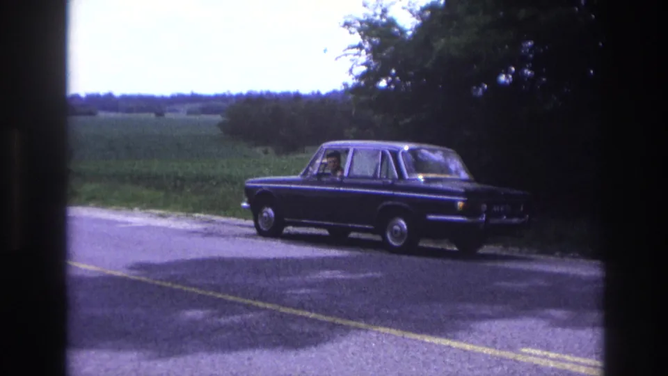 Dark blue 1960s four-door sedan in profile on a paved rural road, likely France, circa 1967; green fields and deciduous trees form the backdrop.