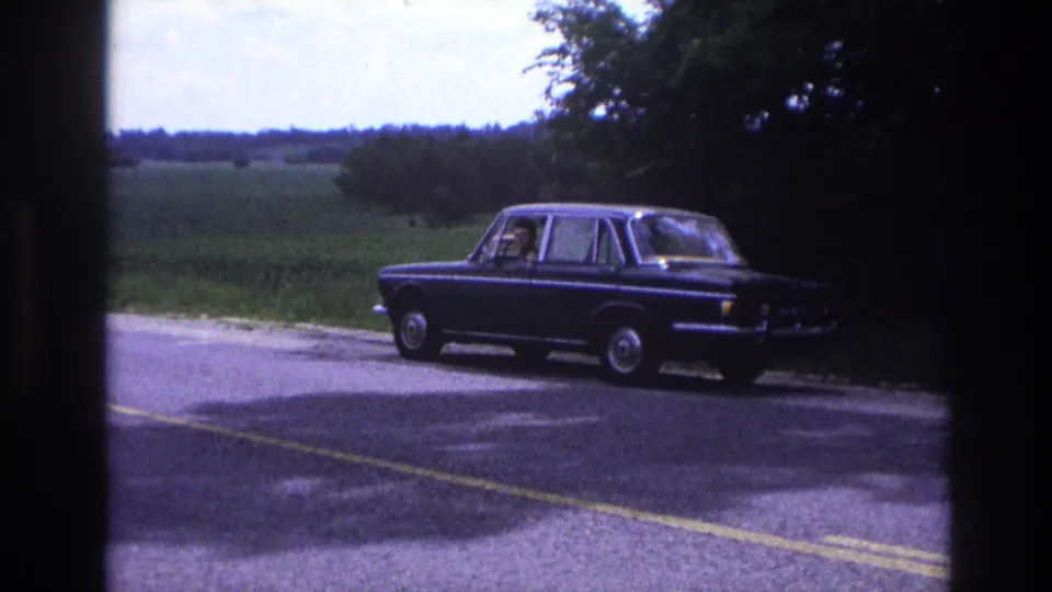 Dark blue 1960s sedan in profile on a French rural road, circa 1967; rolling green farmland and a distant treeline are visible under a pale sky.
