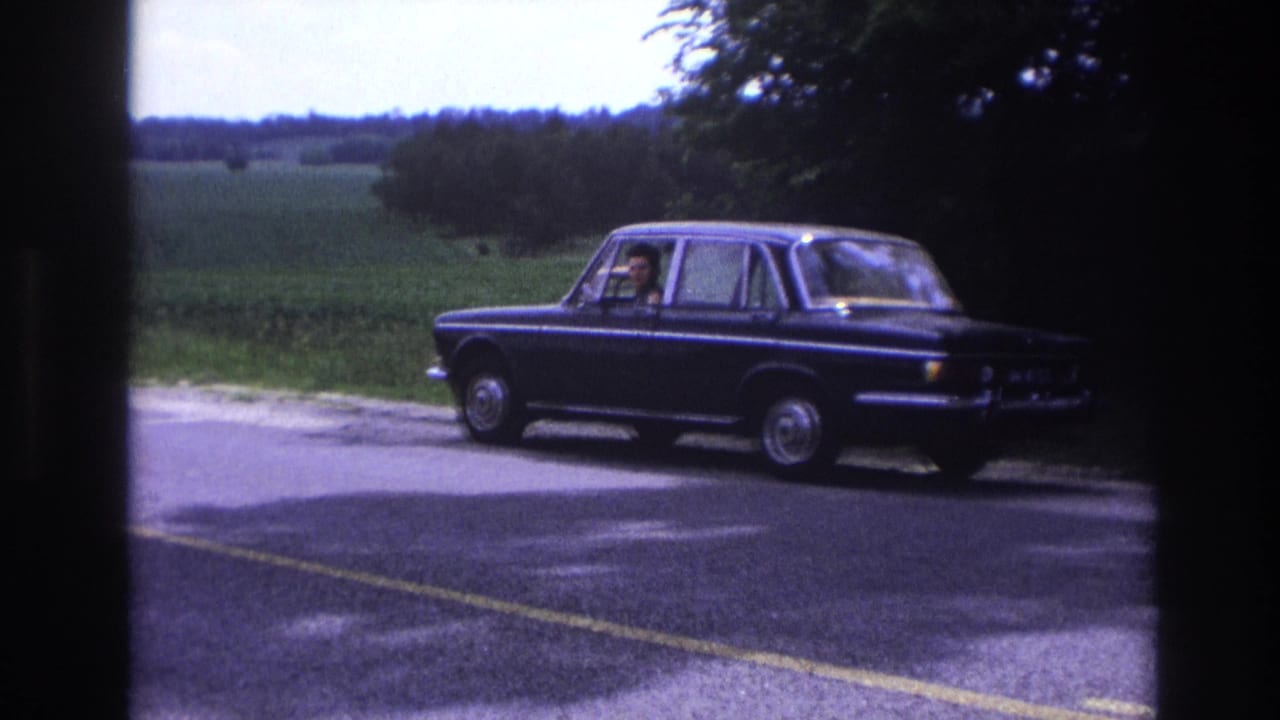 8mm home-movie frame circa 1967, France: dark blue European four-door sedan stopped on a rural road, green summer landscape and treeline behind.