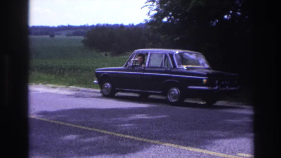 8mm home-movie frame circa 1967, France: dark blue European four-door sedan stopped on a rural road, green summer landscape and treeline behind.