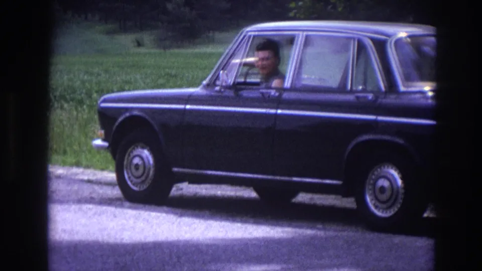 8mm close-up from 1967 France road trip: adult driver's face visible through open window of a dark blue 1960s European sedan stopped roadside, chrome wheel cover prominent.