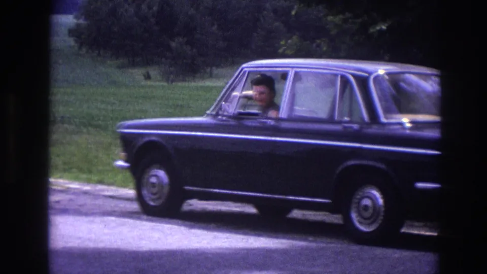 Final 8mm frame of 1967 France road-trip clip: dark blue sedan stopped at rural roadside, driver visible through window, green summer countryside behind.