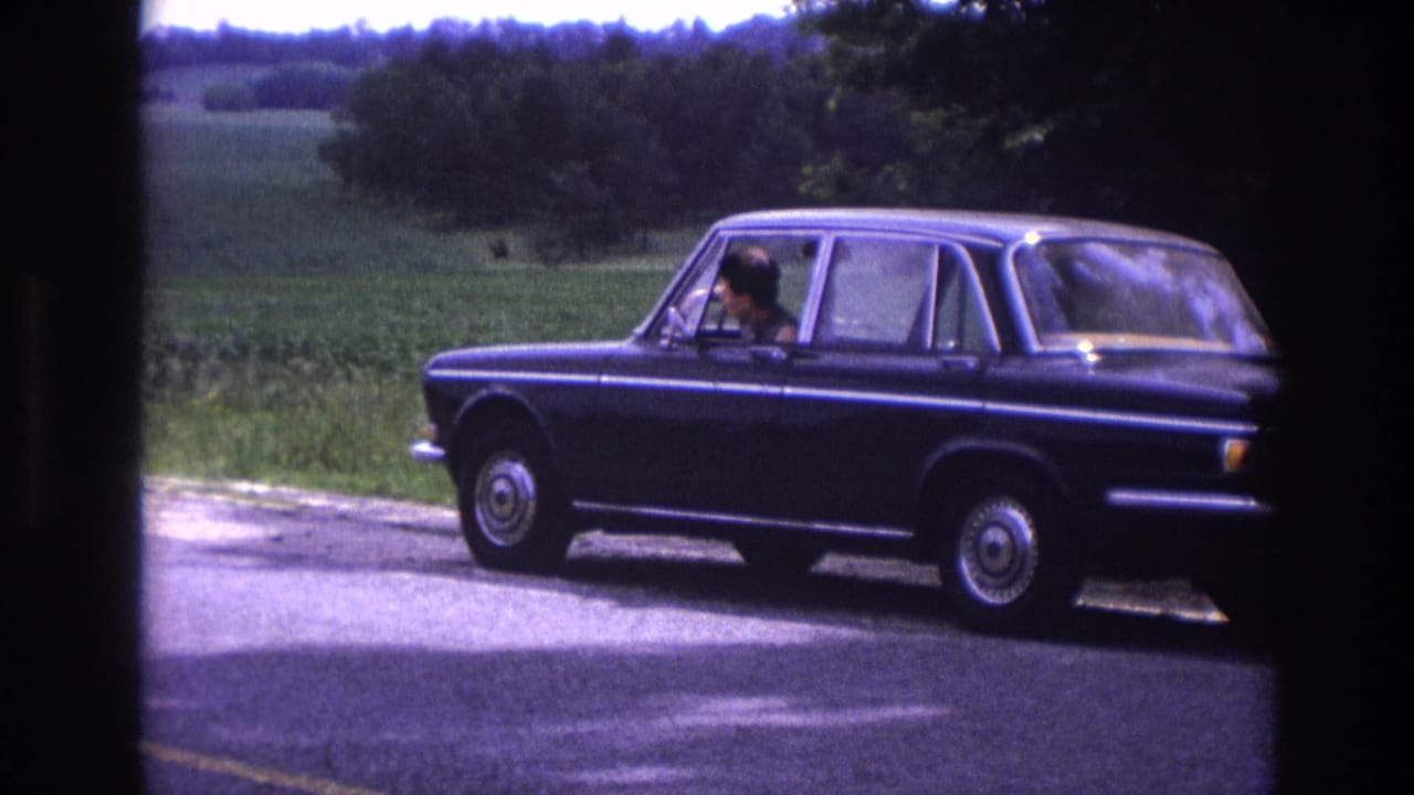 Mid-1960s European four-door sedan stopped on a rural road in France, male driver at the wheel, summer farmland in background, 8mm home movie.