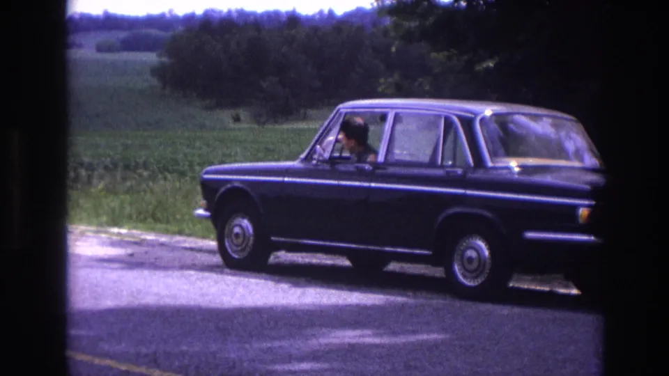 Mid-1960s European four-door sedan stopped on a rural road in France, male driver at the wheel, summer farmland in background, 8mm home movie.