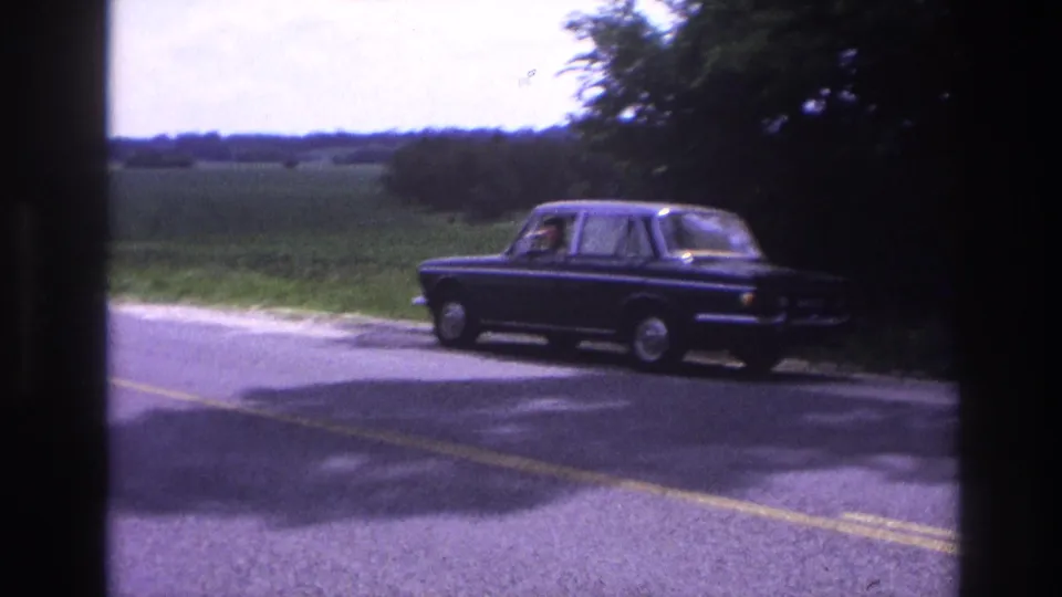 Mid-1960s dark sedan at a bend on a French rural road, male driver visible, summer foliage in background, 8mm film stock.