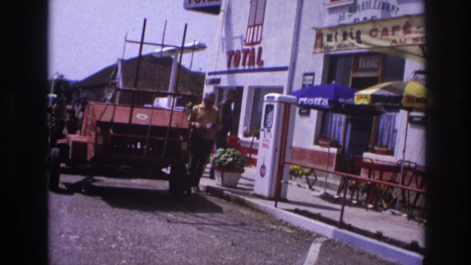 French village roadside stop with visible Total petroleum sign, red agricultural tractor, café with yellow outdoor umbrellas and people nearby, France 1967, 8mm film.