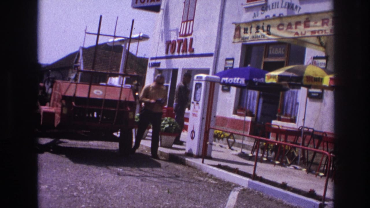 Man in yellow shirt walking toward camera past a Total petrol station; red agricultural trailer at left, café-restaurant sign in background.