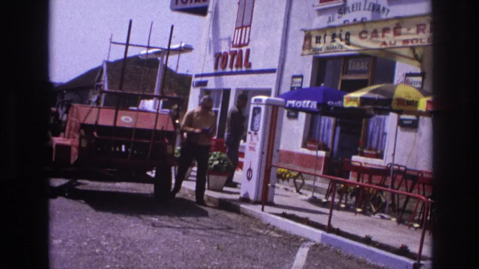 Man in yellow shirt walking past a Total petrol station and café-restaurant in provincial France, circa 1967, captured on 8mm film.