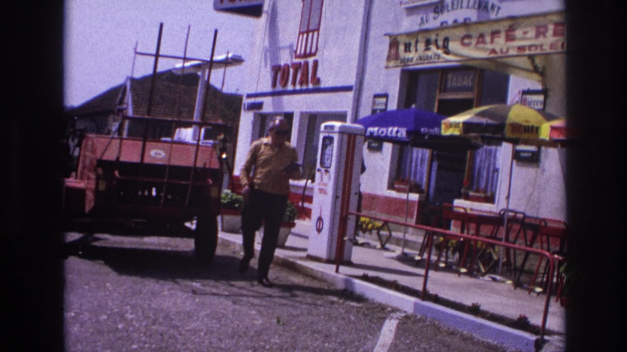 Man in yellow shirt walking past Total petrol station and café in provincial France, circa 1967; second figure visible in background.