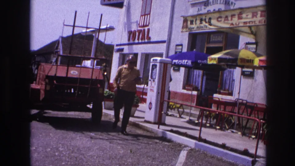 Man in yellow shirt walking past Total petrol station and café in provincial France, circa 1967; second figure visible in background.