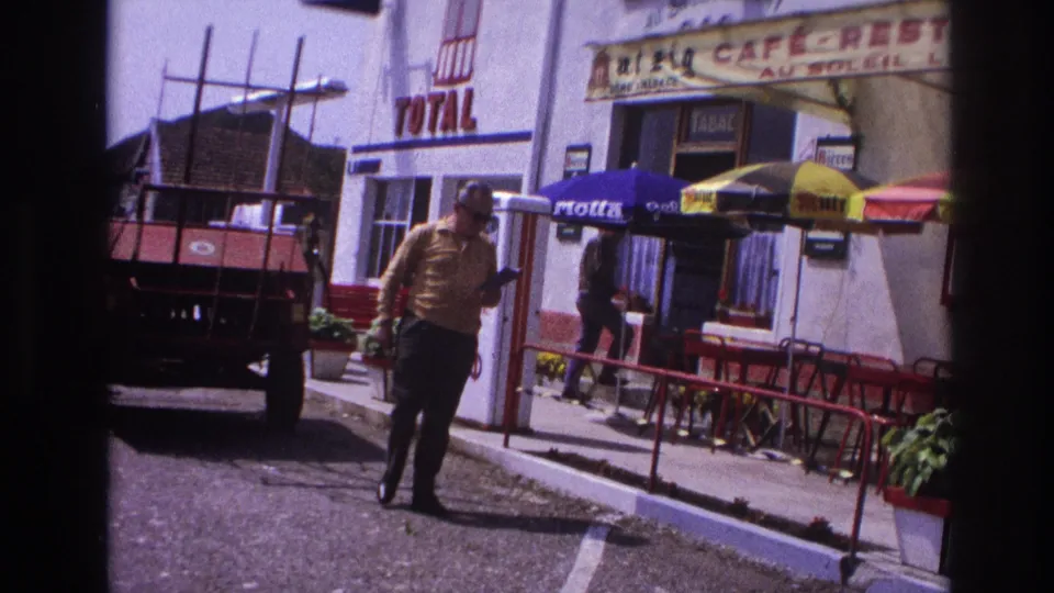 Man in yellow shirt walking in foreground with something in hand, Total petrol station and café-restaurant with umbrellas behind, provincial France, 1967.