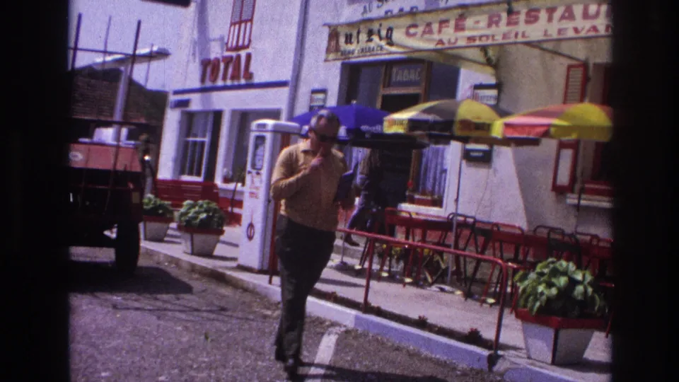 Man in yellow shirt passing close to camera as clip ends, Total petrol station and café-restaurant in background, provincial France, 1967.