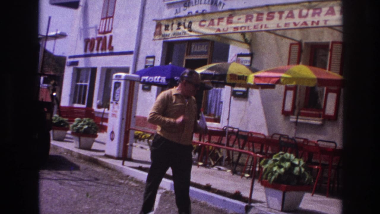 Middle-aged man in mustard long-sleeve shirt and sunglasses walks past Café-Restaurant Au Soleil Levant, France, circa 1967, with Total signage and Motta umbrella visible.