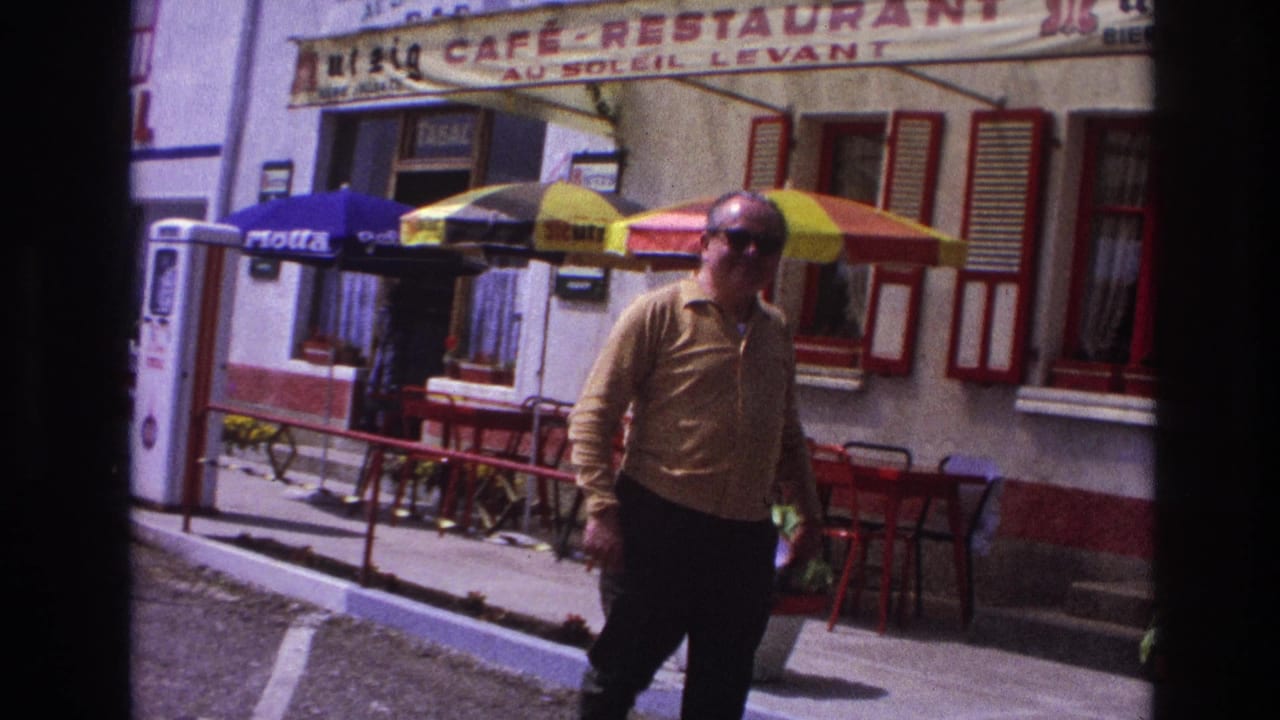 Man in mustard shirt approaching camera past French café with Total pump, Motta umbrella, and red shuttered building.