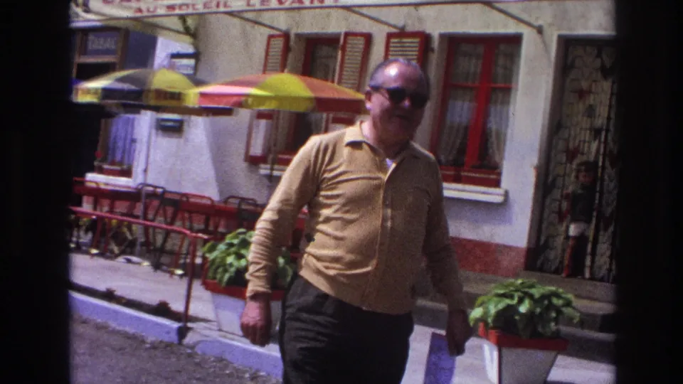 Medium shot of man in mustard long-sleeve shirt and dark sunglasses walking past French café terrace, circa 1967; yellow umbrella and red chairs visible in background.