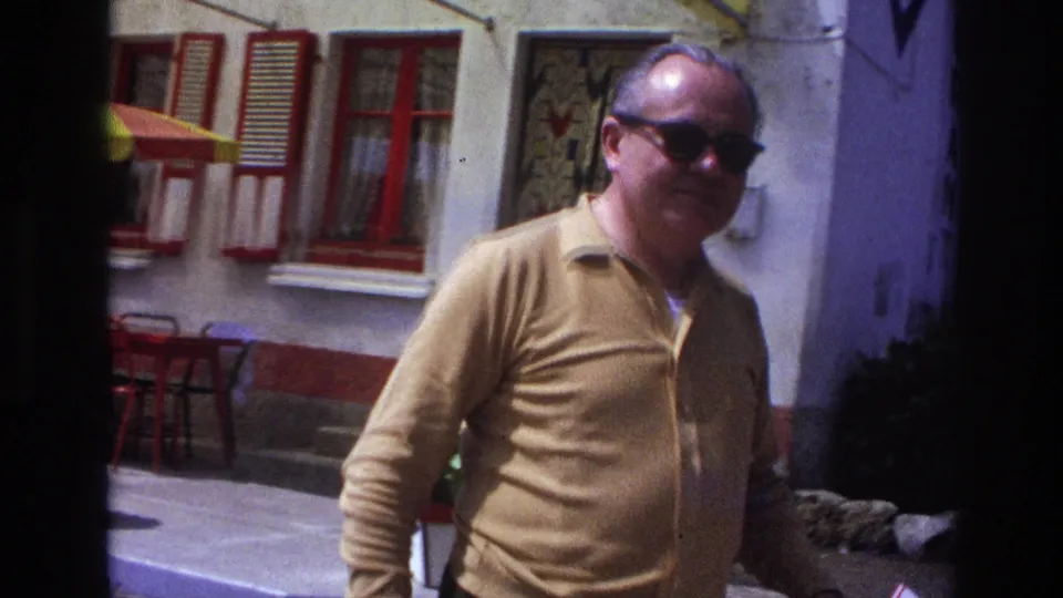 Close shot of middle-aged man in mustard long-sleeve shirt and large dark sunglasses; red-shuttered building and yellow terrace umbrella visible in background, France, circa 1967.