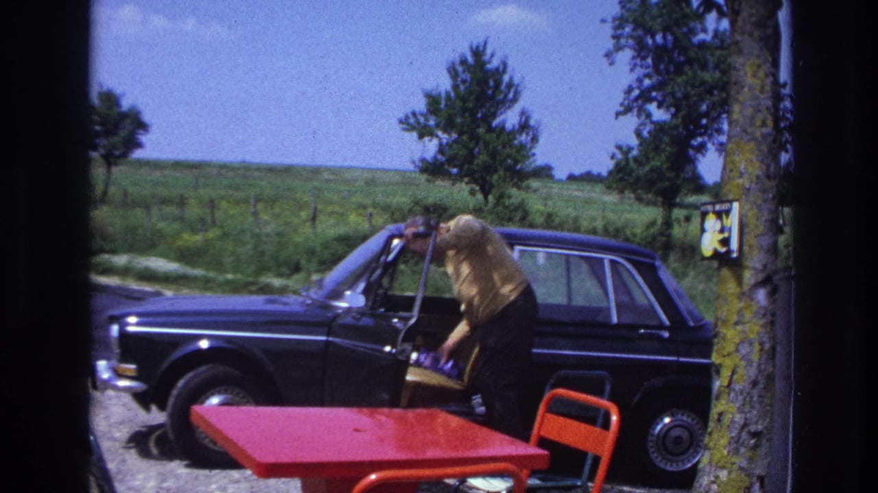 Man in yellow shirt leaning into the open driver's door of a dark late-1960s European sedan at a rural French roadside, red table and orange chair in the foreground.