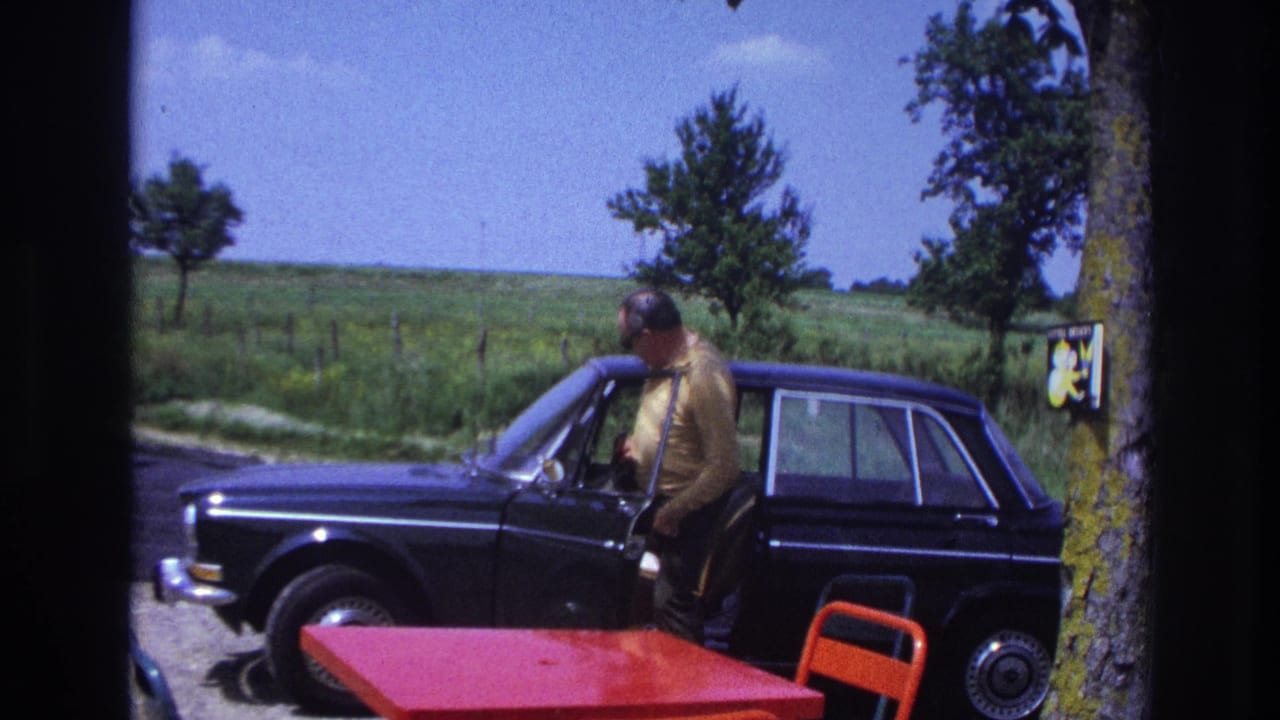 Man in yellow shirt standing beside open driver's door of dark sedan; red table and orange chair visible in foreground.
