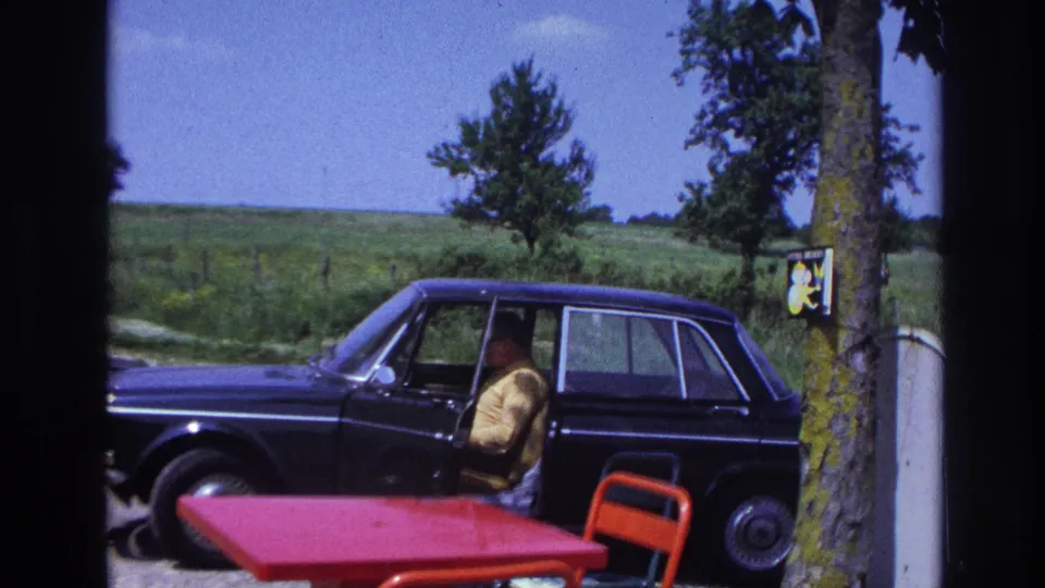 Man in yellow shirt fully settled into the driver's seat of a dark late-1960s European sedan at a rural French roadside rest area, circa 1967.