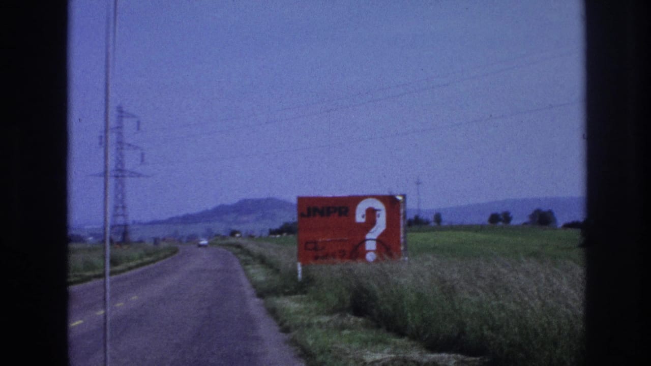 Red roadside billboard reading 'JNPR' with a large white question mark, beside a rural two-lane road with green fields and electricity pylons in the background.