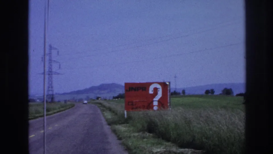 8mm frame from 1967 France: a red billboard marked 'JNPR' with a white question mark stands roadside as a rural road stretches toward low hills under a pale blue sky.