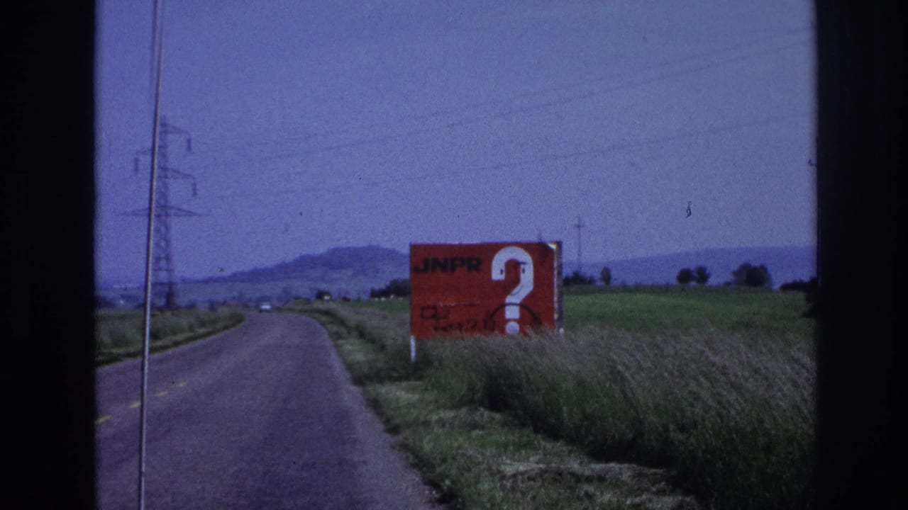 1967 France: red JNPR advertising billboard with oversized question mark, framed by green fields, power pylons, and a low hilly horizon, filmed from a moving vehicle.