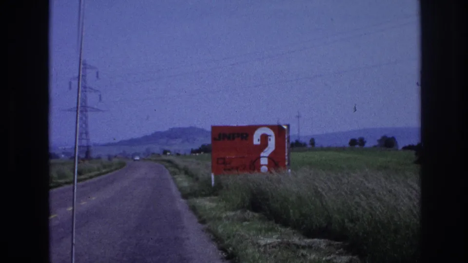 1967 France: red JNPR advertising billboard with oversized question mark, framed by green fields, power pylons, and a low hilly horizon, filmed from a moving vehicle.