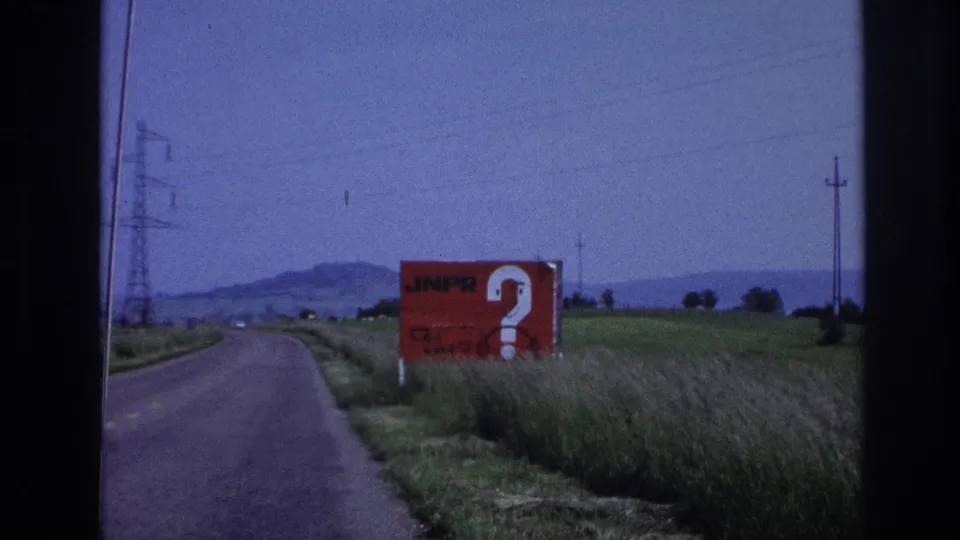 1967 France 8mm: red JNPR billboard with white question mark dominates the roadside, with a utility pole visible mid-distance against hazy hills and a pale sky.