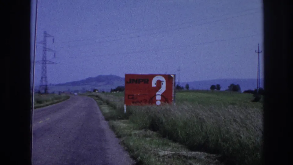 1967 France: red JNPR question-mark billboard passing to the right of frame as the vehicle continues, rural fields, transmission pylons, and low hills in the background.