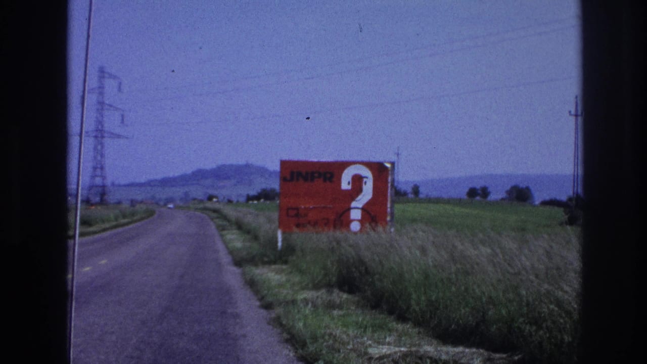 8mm home-movie frame, France c.1967: two-lane rural road with a large red 'JNPR' roadside billboard and white question mark, transmission pylons and low hills in background.
