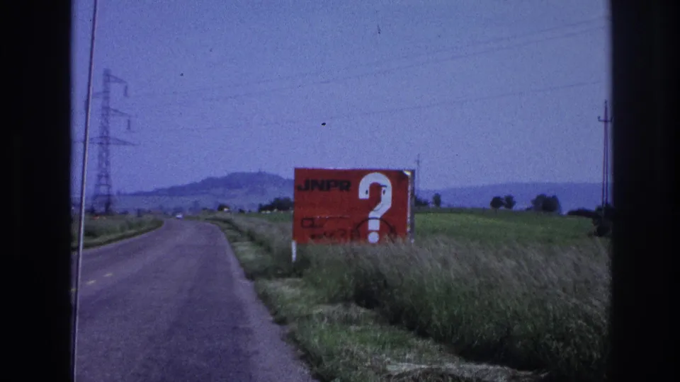 8mm home-movie frame, France c.1967: two-lane rural road with a large red 'JNPR' roadside billboard and white question mark, transmission pylons and low hills in background.