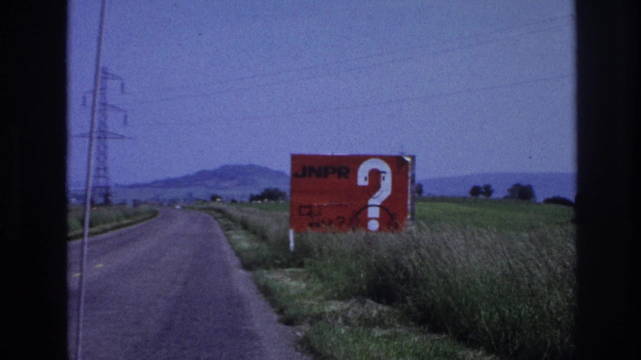 8mm driving shot, France c.1967: road stretching ahead with red 'JNPR?' billboard on right verge, high-tension pylons and low hills visible in the distance.