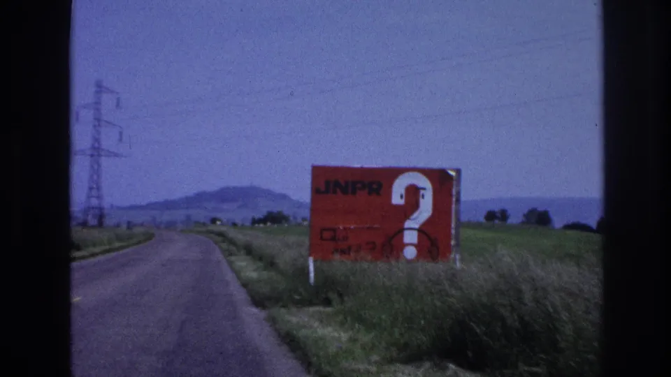 8mm frame, France c.1967: red 'JNPR?' roadside billboard at mid-frame right, green grass verge, high-tension pylons against a hazy low horizon.
