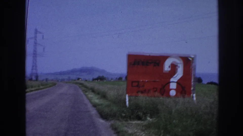 8mm frame, France c.1967: large red 'JNPR' billboard with white question mark fills the right side of frame as the vehicle passes, rural road and pylons receding ahead.